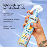 Amika power hour curl refreshing spray held by a hand with blue nail polish against a light blue background.