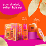 Amika hair care gift set featuring a full-size hair oil bottle, a full-size hair mask jar, and its decorative packaging box, arranged on a bright orange and magenta background with paper-cut style gold accents and text labels.