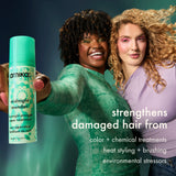 Two women holding a bottle of amika midnight mender hair treatment against a dark background.