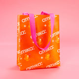 image of an amika premium small tote bag with a logo and sea buckthorn berry print. the bag is a rectangle shopping tote shape with an orange background, white logo and stars, and a pink handle. 