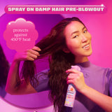 A woman with long, damp hair is shown using Amika's Brooklyn Bombshell Blowout Spray, preparing her hair for a blowout. The scene is bathed in pink and purple lighting, creating a vibrant atmosphere. Text at the top reads 'Spray on Damp Hair Pre-Blowout,' and a pink cloud-shaped graphic on the left highlights that the product 'protects against 450°F heat.' The woman is smiling, holding the spray bottle in one hand and running her other hand through her hair.